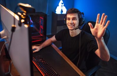 Professional cyber gamer having live stream, waving hand to followers and subscribers of his internet channel, recording vlog via webcam while playing online video game in room. Neon color background